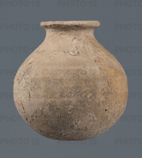 Globular vessel, unknown Northern Mesopotamian workshop, Early Jazira III period ca. 2600–ca. 2350 BC. A globular vessel whose body rises about 3/4 of its height into sloping arms, from which emerges a low, narrow neck terminating in a thickened, flared rim. The transition from body to arms is emphasized by a shallow, wide horizontal groove. Vessels for oils and ointments; vice-free vessel. Al-Jazira Northern Mesopotamia; Khabur basin, Tell Jassa el Gharbi Northeastern Syria. Early Dynastic III period ca. 2600–ca. 2350 BC. Vessel; height 10 cm, width 9.7 cm., A rounded earthen vessel with a narrow neck and flared rim is centered against a solid background. The surface of the vessel is textured and weathered with areas of roughness and small chips. The vessel shows an overall light tan to beige coloration with patches of darker brown and gray mottling, and subtle variations in hue across its body. The rim appears slightly uneven and worn, and the body has a few shallow depressions and i...