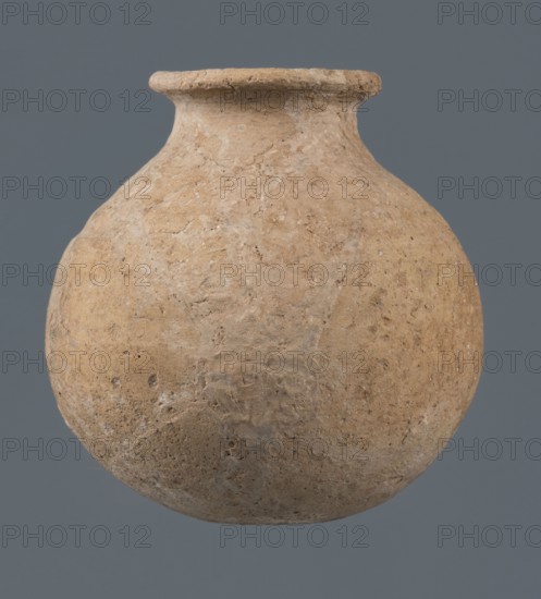 Globular vessel, unknown Northern Mesopotamian workshop, Early Jazira III period ca. 2600–ca. 2350 BC Early Dynastic III period. A globular vessel whose body rises to three-quarters height into sloping arms, from which a low narrow neck arises, terminating in a thickened, flared rim. The transition from body to arms is emphasized by a shallow, wide horizontal groove. Circa 2600–ca. 2350 BC. Origin: Northern Mesopotamia Khabur basin, Tell Jassa el Gharbi, northeastern Syria. Function: vessel for oils and ointments; vice-free vessel. Material/type: vessel; height 10 cm, width 9.7 cm., A rounded, bulbous clay vessel with a short neck and flared rim sits centered against a solid muted gray background; the vessel surface is rough and textured with an uneven, weathered appearance showing a mix of beige, tan, light brown, and pale gray tones.