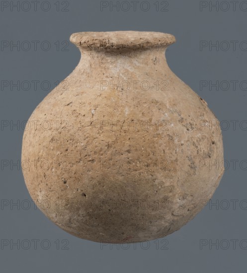 Globular vessel, unknown Northern Mesopotamian workshop, Early Jazira III period ca. 2600–ca. 2350 BC. A globular vessel whose body rises for 3/4 of its height into sloping arms, from which a low, narrow neck emerges, terminating in a thickened, flared rim. The transition from the body to the arms is emphasized by a shallow, wide horizontal groove. c. 2600–ca. 2350 BC; Early Dynastic III period. From Northern Mesopotamia Khabur basin, Tell Jassa el Gharbi Northeastern Syria. Vessel for oils and ointments; vice-free. Height 10 cm, diameter 9.7 cm., A rounded, bulbous ceramic vessel with a short neck and a flared rim sits against a plain background; the vessel's surface is rough and weathered with a textured, cracked appearance showing a range of beige and light brown tones with small patches of darker brown and hints of pale off-white, and the background is a muted gray-blue.