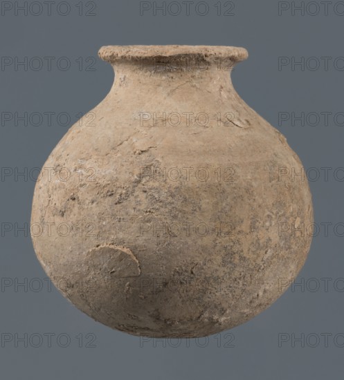Globular vessel, unknown Northern Mesopotamian workshop, Early Jazira III period ca. 2600–ca. 2350 BC. A globular vessel whose body rises to about 3/4 height into sloping arms, from which a low, narrow neck emerges, terminating in a thickened, flared rim. The transition from body to arms is emphasized by a shallow, wide horizontal groove. Early Dynastic III period ca. 2600–ca. 2350 BC. Function: vessel for oils and ointments; vice-free vessel. Origin: Northern Mesopotamia Khabur basin, Tell Jassa el Gharbi, Northeastern Syria. Material: ceramic vessel. Dimensions: height 10 cm, width 9.7 cm., A rounded, weathered clay vessel with a narrow neck and flared rim sits against a solid background. The vessel's surface is rough and uneven with patches of lighter and darker tones in shades of beige, tan, brown, and gray, showing areas of wear, pitting, and small deposits. The rim appears slightly irregular and textured, and the body shows subtle variations in color and texture, including a few ...