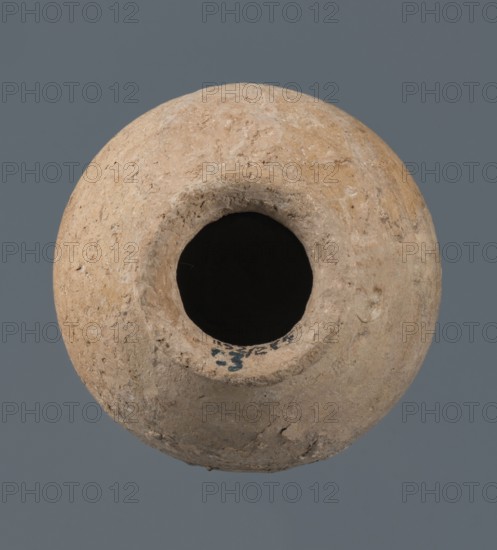 A globular vessel, likely from a Northern Mesopotamian workshop, Early Jazira III / Early Dynastic III period ca. 2600–ca. 2350 BC. The body rises to about 3/4 height into sloping shoulders from which a low, narrow neck emerges, terminating in a thickened, flared rim; the transition from body to shoulders is emphasized by a shallow, wide horizontal groove. Made for oils and ointments; vice-free ceramic. Origin: Al-Jazira Khabur basin, Northern Mesopotamia, Tell Jassa el Gharbi Northeastern Syria. Dimensions: height 10 cm; width 9.7 cm., A round, rough-textured clay vessel with an open circular mouth viewed from above, showing a dark empty interior and uneven, weathered surfaces with small pits and chips; the vessel is in shades of beige, tan, light brown, gray, black inside the opening, and the background is a muted slate gray.