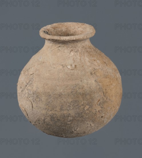 Globular vessel, unknown Northern Mesopotamian workshop, Early Jazira III period ca. 2600–ca. 2350 BC. A globular vessel whose body rises to about 3/4 height into sloping arms, from which a low narrow neck emerges, terminating in a thickened, flared rim. The transition from body to arms is emphasized by a shallow, wide horizontal groove. Early Dynastic III period ca. 2600–ca. 2350 BC. Function: vessel for oils and ointments; vice-free vessel. Al-Jazira Northern Mesopotamia; Khabur basin, Tell Jassa el Gharbi Northeastern Syria, c. 2600–ca. 2350 BC. Material: ceramic vessel; height 10 cm, width 9.7 cm., A rounded, weathered clay vessel with a narrow neck and flared rim sits against a plain gray background; the vessel's surface shows a rough, uneven texture with small chips and worn areas in shades of beige, tan, light brown, and muted gray, and a few darker spots and patches of discoloration.
