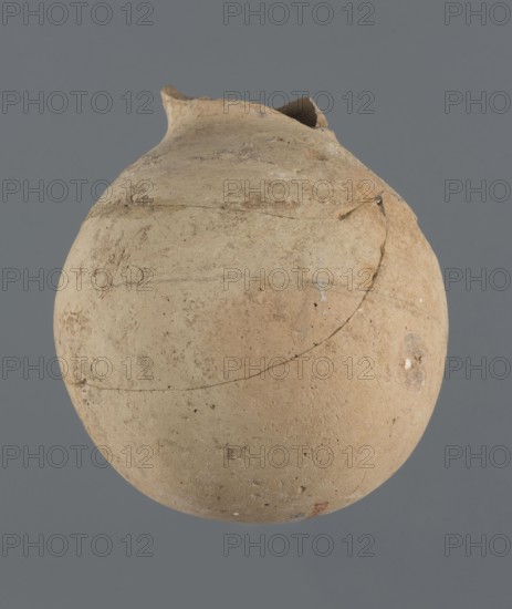 Globular vessel, unknown Northern Mesopotamian workshop Khaburu Basin, vice-free vessel, Early Jazira III period ca. 2600–ca. 2350 BC, vessel for oils and ointments, clay, Early Dynastic III period ca. 2600–ca. 2350 BC, Al-Jazira Syria/Iraq, Mesopotamia, globular vessel with spherical belly merging at 3/4 height into short sloping handles and a narrow slightly flared neck; part of the neck and spout missing. Origin: Northern Mesopotamia Khabur basin, Tell Jassa el Gharbi north-eastern Syria. Vessel, height 13 cm, width 10.9 cm., A rounded, weathered clay vessel with a rough surface in shades of beige, tan, and light brown floats against a plain gray background; the vessel shows irregularities and surface blemishes, a chipped rim with a broken section, visible hairline cracks and repair lines, and a few small darker spots and lighter flecks across its body.