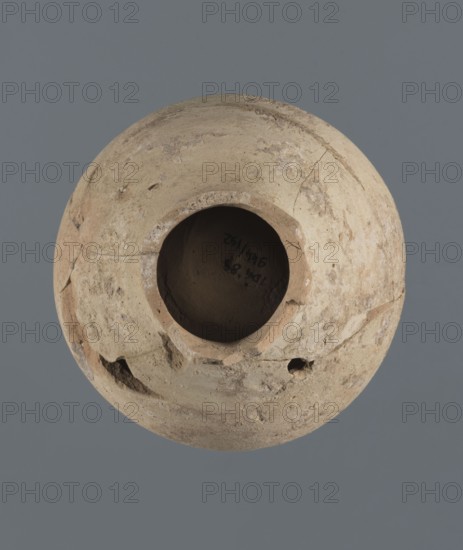 Globular vessel, unknown Northern Mesopotamian workshop Khaburu Basin, vice-free vessel from Early Jazira III period ca. 2600–ca. 2350 BC, used for oils and ointments, clay; Early Dynastic III period ca. 2600–ca. 2350 BC; Al-Jazira Syria/Iraq/Mesopotamia. Vessel with a spherical belly that at three-quarters height merges into short, sloping shoulders with a narrow, slightly flared neck. Part of the neck and spout is missing. Origin: Northern Mesopotamia Khabur basin, Tell Jassa el Gharbi north-eastern Syria. Height: 13 cm; width: 10.9 cm., A round, weathered clay vessel viewed from above with a wide central opening, showing a rough, uneven rim and surface with chips and cracks; the vessel's exterior displays a mix of beige, tan, and light brown tones with patches of darker brown and pale off-white areas; the interior appears darker with shadowed brown and grayish hues; the background is a solid muted gray.