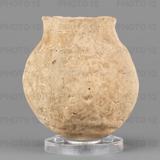 Spherical vessel, unknown Northern Mesopotamian workshop, viseless, hand-made miniature vessel with a rounded bottom and a low neck transitioning into a slightly tilted and undulating short rim with a pointed tip; irregular, wide vertical grooves on the body. 1st half of the 3rd millennium BC; Early Jazira III period ca. 2600–ca. 2350 BC. Tell Jassa el Gharbi North-Eastern Syria; Northern Mesopotamia Khabur basin. Vessel; height 7.9 cm, width 7.1 cm, weight 176 g., A single rounded vessel with a rough, textured surface sits upright on a clear circular stand against a solid gray background; the vessel is an uneven beige to light tan color with small darker flecks and faint variations in tone across its body, showing irregular pits and scratches in the clay and a slightly wavy rim, while the clear stand appears translucent with subtle reflections.