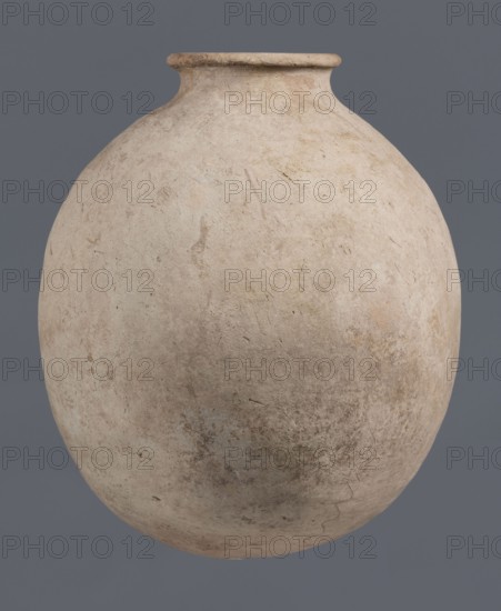Bottle, unknown northern Mesopotamian workshop, Early Jazira III period ca. 2600–ca. 2350 BC / Early Dynastic III period ca. 2600–ca. 2350 BC. Bottle with a bulging, regularly ovoid belly with a rounded bottom, a low narrow neck and a thickened, slightly inclined spout. On the belly, at one-quarter of the vessel’s height, a shallowly engraved potter’s mark in the form of a single sinusoidal-zigzag vertical line. Clay vessel; potter’s marks. Origin: Northern Mesopotamia Khabur Basin, Tell Jassa el Gharbi north-eastern Syria. Height 34.6 cm; width 29.4 cm., A large rounded pottery vessel with a narrow neck and flared rim fills the frame, showing a rough, weathered surface with visible scratches and irregularities; the vessel is primarily shades of beige and pale tan with areas of darker brown and muted gray, and it sits against a solid medium gray background.