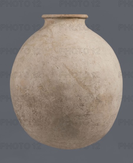 Bottle, unknown northern Mesopotamian workshop, Early Jazira III / Early Dynastic III period ca. 2600–ca. 2350 BC. Clay bottle with a bulging, regularly ovoid belly, rounded bottom, low narrow neck and thickened, slightly inclined spout. On the belly, at one-quarter of the vessel’s height, a shallowly engraved potter’s mark: a single sinusoidal-zigzag vertical line. From Tell Jassa el Gharbi north-eastern Syria, Khabur Basin, northern Mesopotamia. Vessel; height 34.6 cm, width 29.4 cm., A large rounded ceramic vessel with a narrow neck and flared rim dominates the image, showing a weathered surface with uneven texture and small pits and scratches; the vessel is primarily off-white and beige with patches of light tan and subtle grayish tones, set against a solid muted gray background.