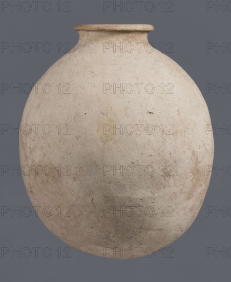 Bottle, unknown northern Mesopotamian workshop, Early Jazira III period ca. 2600–ca. 2350 BC, Early Dynastic III period ca. 2600–ca. 2350 BC. Bottle with a bulging, regularly ovoid belly with a rounded bottom, a low narrow neck and a thickened, slightly inclined spout. On the belly, at one-quarter of the vessel's height, there is a shallowly engraved potter's mark in the form of a single sinusoidal-zigzag vertical line. Material: clay. Origin: Al-Jazira / Khabur Basin, northern Mesopotamia Tell Jassa el Gharbi, north-eastern Syria. Vessel; height 34.6 cm, width 29.4 cm., A large rounded ceramic vessel with a narrow neck and flared rim dominates the image, showing a textured surface with a weathered, matte finish; the vessel is mainly off-white to beige with areas of light tan and pale brown discoloration, along with small dark flecks and faint smudges; the background is a solid medium gray.