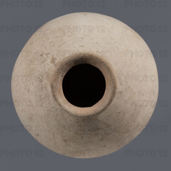 Bottle, unknown northern Mesopotamian workshop, Early Jazira III period ca. 2600–ca. 2350 BC. Bottle with a bulging, regularly ovoid belly, rounded bottom, low narrow neck and thickened, slightly inclined spout. On the belly, at one-quarter of the vessel’s height, a shallowly engraved potter’s mark in the form of a single sinusoidal–zigzag vertical line. Clay vessel; potter’s marks; Early Dynastic III period ca. 2600–ca. 2350 BC. Origin: Al-Jazira Khabur Basin, northern Mesopotamia; findspot Tell Jassa el Gharbi north-eastern Syria. Height 34.6 cm, width 29.4 cm., A round, weathered ceramic vessel viewed from above with a wide circular opening leading into a dark interior, the vessel surface showing a rough, matte texture with small scratches and irregularities; the body is mostly pale beige with areas of warmer tan and faint grayish patches, and the background is a flat medium gray.