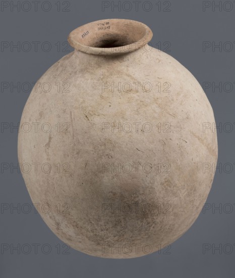 Bottle, unknown northern Mesopotamian workshop, Early Jazira III period ca. 2600–ca. 2350 BC. Clay bottle with a bulging, regularly ovoid belly, rounded bottom, low narrow neck and thickened, slightly inclined spout. On the belly, at one-quarter height, a shallowly engraved potter’s mark in the form of a single sinusoidal-zigzag vertical line. Early Dynastic III period ca. 2600–ca. 2350 BC. Found at Tell Jassa el Gharbi north-eastern Syria; origin: Northern Mesopotamia Khabur Basin. Vessel; height 34.6 cm, width 29.4 cm., A large rounded ceramic vessel with a narrow flared neck and rounded rim is centered against a plain background. The vessel surface appears textured and worn with irregular marks and scratches. The colors visible include various shades of beige, tan, cream, and light brown, with areas of darker brown and muted grayish tones near the lower portion. The rim shows a slightly darker beige and the interior visible at the opening is a deeper brownish shade. The background i...