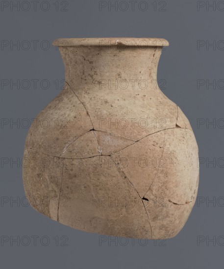 Fragment of a bottle upper 3/4, unknown Northern Mesopotamian workshop, Early Jazira III period ca. 2600–ca. 2350 BC / Early Dynastic III period ca. 2600–ca. 2350 BC. The upper 3/4 of a vice-free bottle with a slender ovoid belly and a high neck terminating in a thickened, outwardly turned rim of triangular cross-section. Materials: clay; vessel. Origin/place of discovery: Tell Jassa el Gharbi north-eastern Syria, Northern Mesopotamia Khabur basin. Dimensions: height 18 cm, width 16 cm., A rounded clay vessel with a wide flared rim sits against a plain gray background; the vessel shows a textured, weathered surface with visible cracks and repairs, and its colors range from pale beige to warm tan and muted brown, with some areas of slightly darker brown and faint reddish tones.