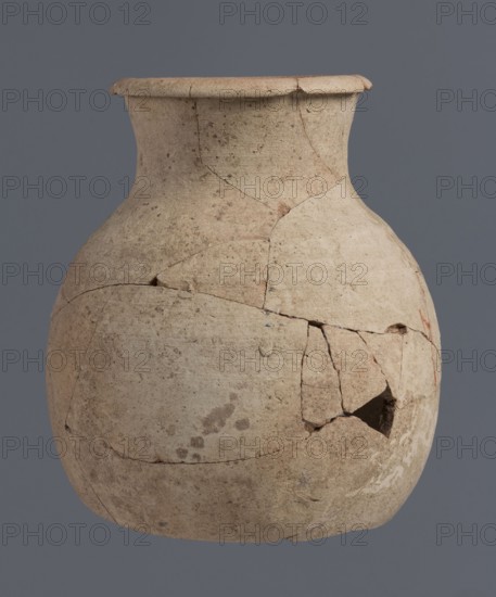 Fragment of a bottle upper 3/4, unknown Northern Mesopotamian workshop; Early Jazira III period ca. 2600–ca. 2350 BC / Early Dynastic III period; vice-free clay bottle with a slender ovoid belly and a high neck terminating in a thickened, outwardly turned rim of triangular cross-section; provenance: Tell Jassa el Gharbi north-eastern Syria, Northern Mesopotamia Khabur basin; vessel; height 18 cm, width 16 cm., A weathered, rounded ceramic jar with a narrow neck and flared rim sits against a plain gray background; the jar's surface is uneven and cracked with visible repairs and missing fragments, showing a muted palette of beige, tan, light brown, and small darker speckles, along with a few faint darker stains and subtle reddish areas near a damaged section.