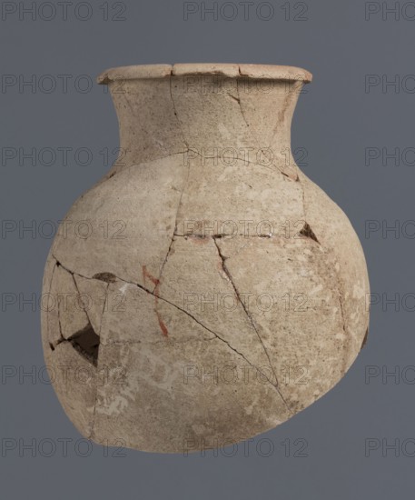 Fragment of a bottle upper 3/4, unknown Northern Mesopotamian workshop; Early Jazira III period ca. 2600–ca. 2350 BC / Early Dynastic III period; vice-free clay vessel bottle. The upper 3/4 of a vice-free bottle with a slender ovoid belly and a high neck terminating in a thickened, outwardly turned rim of triangular cross-section. Origin: Northern Mesopotamia Khabur basin, Tell Jassa el Gharbi north-eastern Syria. Dimensions: height 18 cm, width 16 cm., A rounded ceramic vessel with a wide flared rim, shown against a solid gray background, displaying a textured surface in shades of beige and light tan with darker brown and faint reddish areas; the vessel has visible cracks and repaired joins, some missing fragments creating small gaps, and a few smudged pale markings on the lower area.