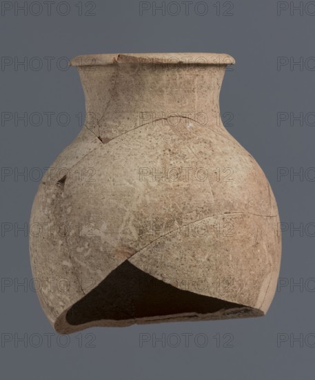 Fragment of a bottle upper 3/4, unknown Northern Mesopotamian workshop, Early Jazira III period ca. 2600–ca. 2350 BC Early Dynastic III period. The upper 3/4 of a vice-free bottle with a slender ovoid belly and a high neck terminating in a thickened, outwardly turned-out rim of triangular cross-section. Clay vessel bottle. Provenance: Tell Jassa el Gharbi north-eastern Syria, Northern Mesopotamia Khabur basin. Height 18 cm; width 16 cm., A weathered, rounded clay vessel with a flared rim and a narrow neck sits against a plain gray background, showing a surface of beige and light brown tones with patches of darker brown and pale speckling; the vessel displays several visible cracks and areas where pieces are missing, revealing an inner, darker interior, and the exterior texture appears rough and porous with irregular shading and subtle worn marks.