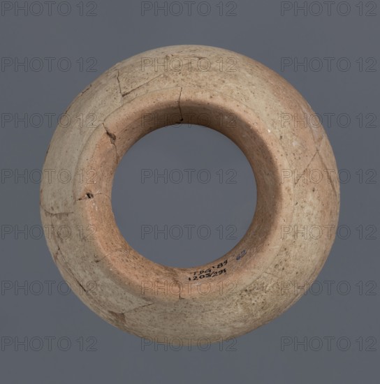 Fragment of a bottle upper 3/4, unknown Northern Mesopotamian workshop, Early Jazira III period ca. 2600–ca. 2350 BC Early Dynastic III period. The upper three-quarters of a vice-free bottle with a slender ovoid belly and a high neck terminating in a thickened, outwardly turned rim of triangular cross-section. Clay vessel, height 18 cm, width 16 cm. Provenance: Tell Jassa el Gharbi north-eastern Syria, Northern Mesopotamia Khabur basin., A round, hollow ceramic vessel shown from above with a wide circular opening and a thick rounded rim, displaying a weathered, slightly uneven surface with visible cracks and repaired joins. The vessel's colors include beige, tan, light brown, and hints of gray, with darker brown speckles and small areas of discoloration. The background is a solid mid-gray color.