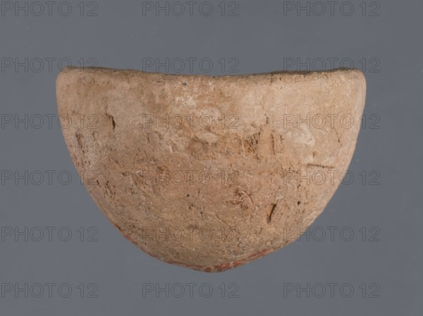 Miniature bowl, unknown Northern Mesopotamian workshop, Early Jazira III period ca. 2600–ca. 2350 BC / Early Dynastic III period ca. 2600–ca. 2350 BC. Clay vessel, miniature bowl with a narrow rounded and missing bottom and a straight rim; arched walls terminating in a straight rim. Origin: Northern Mesopotamia Khabur basin, Tell Jassa el Gharbi North-Eastern Syria. Height 3 cm; width 5 cm., A roughly hemispherical object with a textured, worn surface in shades of tan and light brown set against a flat bluish-gray background; the object shows irregular pits and scratches and a few darker specks, with slightly darker brown areas and faint reddish traces along the lower edge.