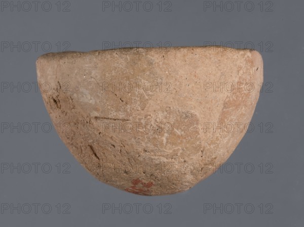 Bowl, unknown Northern Mesopotamian workshop; Early Jazira III period ca. 2600–ca. 2350 BC / Early Dynastic III period ca. 2600–ca. 2350 BC. Miniature bowl with a narrow rounded and missing bottom and a straight rim; arched walls terminating in a straight rim. Clay vessel; miniature vessel. Origin: Northern Mesopotamia Khabur basin, Al-Jazira Syria/Iraq. Found at Tell Jassa el Gharbi north-eastern Syria. Height: 3 cm; Width: 5 cm., A roughly hemispherical clay bowl with a coarse, weathered surface in shades of tan, light brown, and beige, showing patches of darker brown and faint reddish discoloration near the lower edge; the bowl has an uneven rim and visible pits, scratches, and textured irregularities across its exterior, set against a uniform gray background.