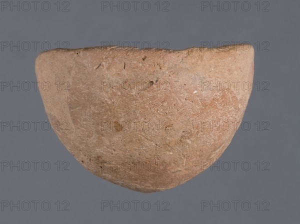 Bowl, unknown Northern Mesopotamian workshop, Early Jazira III period ca. 2600–ca. 2350 BC. Miniature bowl with a narrow rounded and missing bottom and a straight rim; arched walls terminating in a straight rim. Clay vessel, miniature, height 3 cm, width 5 cm. Origin: Northern Mesopotamia Khabur basin, Tell Jassa el Gharbi North-Eastern Syria, Al-Jazira region. Early Dynastic III period, c. 2600–ca. 2350 BC., A roughly hemispherical object with a textured, weathered surface in shades of light brown, tan, and reddish-brown sits against a flat gray background, showing irregular edges, small pits and scratches, and subtle variations in color and surface roughness across its curved exterior.