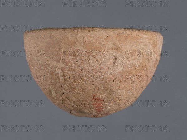 Miniature bowl, unknown Northern Mesopotamian workshop, Early Jazira III period ca. 2600–ca. 2350 BC. Clay; miniature vessel with a narrow rounded missing bottom and a straight rim; arched walls terminating in a straight rim. Early Dynastic III period ca. 2600–ca. 2350 BC. Origin: Northern Mesopotamia Khabur basin, Tell Jassa el Gharbi North-Eastern Syria. Height 3 cm; width 5 cm., A roughly hemispherical bowl with a textured, worn surface in shades of tan, beige, and light brown is centered against a solid gray background; the bowl shows areas of roughness and small pits, with subtle variations of cream and ochre on its exterior and a small area of muted red near the lower rim.