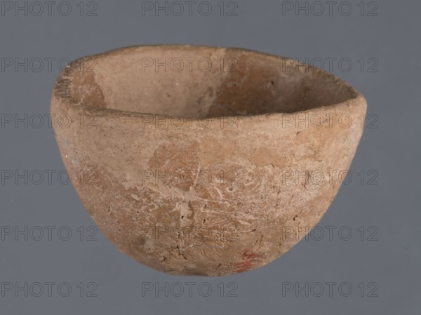 Bowl, unknown Northern Mesopotamian workshop; Early Jazira III period ca. 2600–ca. 2350 BC / Early Dynastic III period ca. 2600–ca. 2350 BC. Miniature bowl of clay with a narrow rounded missing bottom, arched walls terminating in a straight rim, and a straight rim overall. Miniature vessel; height 3 cm, width 5 cm. Origin: Northern Mesopotamia Khabur basin, Tell Jassa el Gharbi North-eastern Syria., A roughly rounded clay bowl with an uneven rim sits against a plain gray background, showing a surface texture that is coarse and weathered with patches of lighter and darker brown, tan, and beige, and a small area of muted red near the lower edge; the interior shows similar earthy tones and worn surfaces, and the overall shape is slightly asymmetrical.