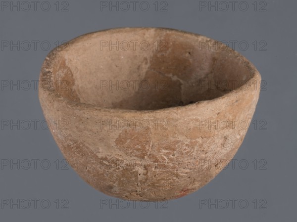 Miniature bowl, unknown Northern Mesopotamian workshop. Early Jazira III / Early Dynastic III period, c. 2600–ca. 2350 BC. Clay vessel; miniature bowl with a narrow rounded missing bottom, arched walls terminating in a straight rim. From Tell Jassa el Gharbi, North-eastern Syria Khabur basin, Northern Mesopotamia. Height 3 cm; width 5 cm., A roughly hemispherical earthenware bowl with a textured, uneven rim and interior surface, showing areas of wear and small pits; the bowl appears in shades of tan, beige, and light brown with slightly darker brown patches and speckles, set against a solid medium gray background.
