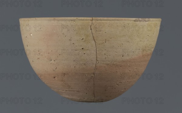 Bowl, unknown Northern Mesopotamian workshop. Early Jazira III period ca. 2600–ca. 2350 BC / Early Dynastic III period. Small round flat-bottomed bowl: walls tilted in the lower half, straight in the upper half, ending with a low, slightly distinct, slightly thickened and rounded rim. Pinkish burn mark visible on the lower part from contact with another bowl. Material: clay. Origin: Northern Mesopotamia Al-Jazira/Khabur Basin, found at Tell Jassa el Gharbi Northeastern Syria. Function: vessel/table bowl. Dimensions: height 7.7 cm; width 12.3 cm; weight 170 g., A rounded ceramic bowl shown against a solid dark gray background, with a visible vertical crack running from rim to base and a surface pitted with small holes and irregularities; the bowl displays a gradient of colors including beige, tan, light brown, pale pinkish tones, and hints of yellow, with subtle horizontal striations and areas of darker brown near the lower portion.