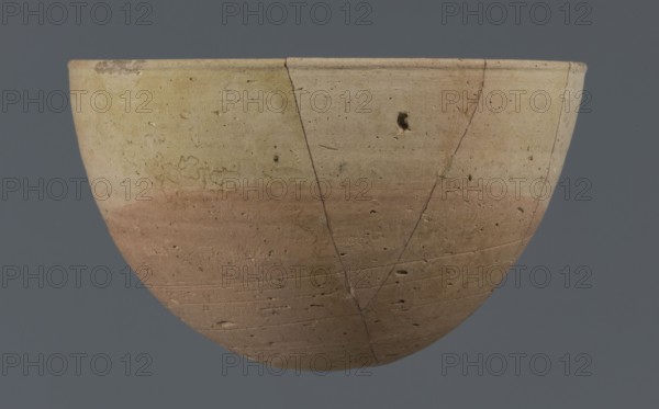 Bowl, unknown Northern Mesopotamian workshop. Early Jazira III period ca. 2600–ca. 2350 BC / Early Dynastic III period ca. 2600–ca. 2350 BC. Clay vessel, table vessel; vice-free. Bowl with a small round flat bottom; walls tilted in the lower half, straight in the upper half, ending with a low, slightly distinct, slightly thickened and rounded rim. Pinkish burn mark in the lower part from contact with the bowl in which it was placed. Ca. 2600–2350 BC. Findspot: Tell Jassa el Gharbi Northeastern Syria, Northern Mesopotamia Khabur Basin. Height 7.7 cm; width 12.3 cm; weight 170 g., A shallow bowl shape with a rounded base and flared rim sits against a solid gray background; the object shows a smooth, worn surface with a gradient of muted colors including beige, pale yellow, light tan, and a washed reddish-brown band across the lower half, and there are visible hairline cracks, small pitted holes, and irregular surface marks across the exterior.