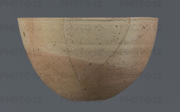 Bowl, unknown Northern Mesopotamian workshop. Bowl with a small round flat bottom. The bowl walls are tilted in the lower half, straight in the upper half, ending with a low, slightly distinct, slightly thickened and rounded rim. In the lower part of the bowl, a pinkish burn mark is visible, left by the bowl in which it was placed. Clay vessel, table vessel; vice-free vessel. Early Jazira III period ca. 2600–ca. 2350 BC / Early Dynastic III period ca. 2600–ca. 2350 BC. Al-Jazira Northern Mesopotamia; Khabur Basin, Tell Jassa el Gharbi Northeastern Syria, ca. 2600–ca. 2350 BC. Height 7.7 cm; width 12.3 cm; weight 170 g., A shallow rounded bowl viewed from the side against a gray background, showing a weathered surface with a gradient of beige to light brown and hints of pink, small dark speckles and pits across the surface, several visible hairline cracks and lines forming irregular patterns, and subtle horizontal striations and texture variations on the clay.
