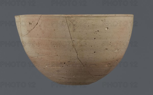 Bowl, unknown Northern Mesopotamian workshop, bowl with a small round flat bottom. The bowl walls are tilted in the lower half, straight in the upper half, ending with a low, slightly distinct, slightly thickened and rounded rim. In the lower part of the bowl, a pinkish burn mark is visible, left by the bowl in which it was placed. Clay vessel, table vessel; vice-free. Early Jazira III period / Early Dynastic III period, ca. 2600–ca. 2350 BC. Findspot: Tell Jassa el Gharbi Northeastern Syria; origin: Northern Mesopotamia Khabur Basin, Al-Jazira region. Height 7.7 cm; width 12.3 cm; weight 170 g., A rounded bowl with a smooth rim sits against a flat background, showing an outer surface marked by several hairline cracks and small pits. The bowl's finish displays horizontal tool marks and subtle variations in color, with areas of beige, light tan, muted pink, and pale brown blending across its curved surface. The background is a uniform dark gray.