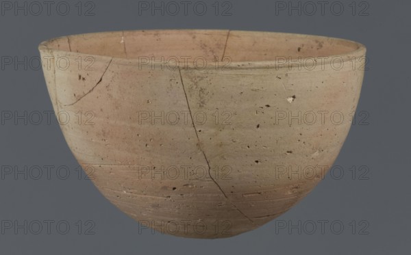 Bowl, unknown Northern Mesopotamian workshop. Early Jazira III period ca. 2600–ca. 2350 BC / Early Dynastic III period ca. 2600–ca. 2350 BC. Clay table vessel with a small round flat bottom; walls tilted in the lower half, straight in the upper half, ending with a low, slightly distinct, slightly thickened and rounded rim. Pinkish burn mark visible on the lower part from contact with the bowl in which it was placed. Origin: Al-Jazira Khabur Basin, Northern Mesopotamia; found at Tell Jassa el Gharbi Northeastern Syria. Height 7.7 cm, width 12.3 cm, weight 170 g., A rounded ceramic bowl with a wide rim, showing visible hairline cracks and surface pits, in shades of beige, tan, light brown, and faint pink, set against a solid muted gray background.