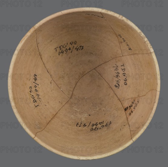Bowl, unknown Northern Mesopotamian workshop, bowl with a small round flat bottom. The bowl walls are tilted in the lower half, straight in the upper half, ending with a low, slightly distinct, slightly thickened and rounded rim. In the lower part of the bowl, a pinkish burn mark is visible, left by the bowl in which it was placed. Clay vessel, table vessel, vice-free, ca. 2600–2350 BC; Early Jazira III / Early Dynastic III period. Found at Tell Jassa el Gharbi Northeastern Syria; origin Northern Mesopotamia Khabur Basin. Height 7.7 cm, width 12.3 cm, weight 170 g., A round, shallow ceramic bowl viewed from above with an interior surface in shades of tan and beige showing a slightly darker central area and lighter rim; the bowl has visible cracks and a few repaired seams forming a patchwork of segments, with some small spots of discoloration and surface irregularities; the background is a flat gray.