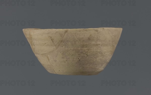 Bowl, unknown Northern Mesopotamian workshop, Early Jazira III period ca. 2600–ca. 2350 BC. Clay table vessel with a flat round bottom showing traces of trimming. Straight, flaring conical walls terminating in a straight, non-distinctive rim; the whole is asymmetrical. Circa 2600–2350 BC. Tell Jassa el Gharbi north-eastern Syria, Northern Mesopotamia Khabur Basin., A shallow, wide bowl with an uneven rim and a rounded base, showing a rough, textured surface with visible tool marks and small pits; the bowl appears in shades of beige and light brown with subtle variations and patches of darker brown, set against a solid dark gray background.