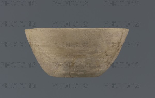 Bowl, unknown Northern Mesopotamian workshop, Early Jazira III period ca. 2600–ca. 2350 BC / Early Dynastic III period ca. 2600–ca. 2350 BC. Clay table vessel; bowl with a flat round bottom with traces of trimming; straight, flaring conical walls terminating in a straight, non-distinctive rim. The whole is asymmetrical. c. 2600–c. 2350 BC; Tell Jassa el Gharbi North-Eastern Syria, Northern Mesopotamia Khabur Basin., A roughly bowl-shaped object with a matte, textured surface sits against a solid background; the object is primarily beige to light brown with areas of darker brown and faint grayish patches, showing subtle horizontal striations and small pits on its surface, and a few visible hairline cracks and irregularities along its rim and lower portion, while the background is a muted charcoal to slate gray.