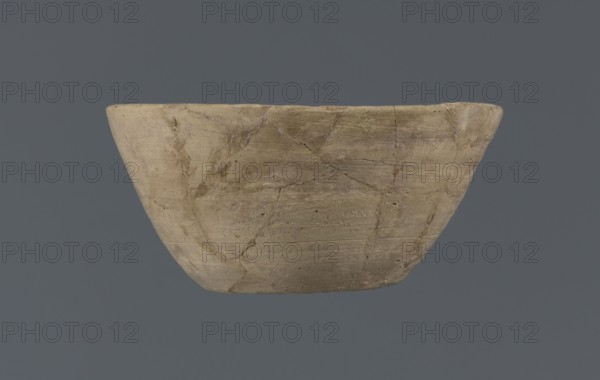 Bowl, unknown Northern Mesopotamian workshop, Early Jazira III period ca. 2600–ca. 2350 BC. Clay table vessel: bowl with a flat round bottom with traces of trimming, straight flaring conical walls terminating in a straight, non-distinctive rim; whole is asymmetrical. Provenance: Northern Mesopotamia Khabur Basin, Al-Jazira; Tell Jassa el Gharbi North-Eastern Syria., A shallow, rounded bowl with a wide rim shown against a solid muted blue-gray background; the bowl has an uneven, textured surface with visible cracks and repair lines and appears in several shades of beige, tan, and light brown with subtle darker streaks and spots, showing a matte, slightly rough finish.