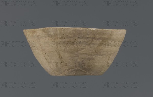 Bowl, unknown Northern Mesopotamian workshop, Early Jazira III period ca. 2600–ca. 2350 BC. Clay table vessel — bowl with a flat round bottom with traces of trimming; straight, flaring conical walls terminating in a straight, non-distinctive rim. The whole is asymmetrical. c. 2600–c. 2350 BC; Early Jazira III period. Tell Jassa el Gharbi North-Eastern Syria; Northern Mesopotamia Khabur Basin., A shallow, wide bowl with an irregular rim displayed against a solid dark gray background, the bowl showing a rough, weathered surface with visible cracks and repair lines; the bowl's color is a mix of beige, light brown, and tan with subtle darker brown streaks and patches, and there are small areas of pale off-white along the rim.