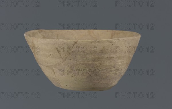 Bowl, unknown Northern Mesopotamian workshop; Early Jazira III period ca. 2600–ca. 2350 BC; Early Dynastic III period ca. 2600–ca. 2350 BC. Clay table vessel bowl with a flat round bottom showing traces of trimming. Straight, flaring conical walls terminating in a straight, non-distinctive rim. The whole is asymmetrical. c. 2600–c. 2350 BC; found at Tell Jassa el Gharbi north-eastern Syria, Northern Mesopotamia Khabur Basin., A roughly rounded, shallow bowl with an uneven rim sits against a flat gray background; the bowl shows a textured, matte surface in shades of beige, tan, and light brown with subtle areas of pale greenish and grayish discoloration, small pits and scratches, and visible horizontal tool marks and faint linear impressions on its exterior.