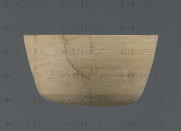 Bowl, unknown northern Mesopotamian workshop; Early Jazira III period ca. 2600–ca. 2350 BC; clay vessel; bowl/table vessel with flat oval bottom from which straight, slightly tilted walls rise in a short arc, terminating in a straight, not distinct rim; Early Dynastic III period ca. 2600–ca. 2350 BC; Al-Jazira Northern Mesopotamia, Khabur basin; found at Tell Jassa el Gharbi north-eastern Syria; height 7 cm, width 12.2 cm., A shallow, rounded container with a smooth, matte surface in pale beige and light tan tones, showing a few darker tan streaks and subtle grayish lines; the vessel has visible horizontal striations and a couple of thin, curved cracks with slightly darker edges; the background is a solid muted slate-gray.