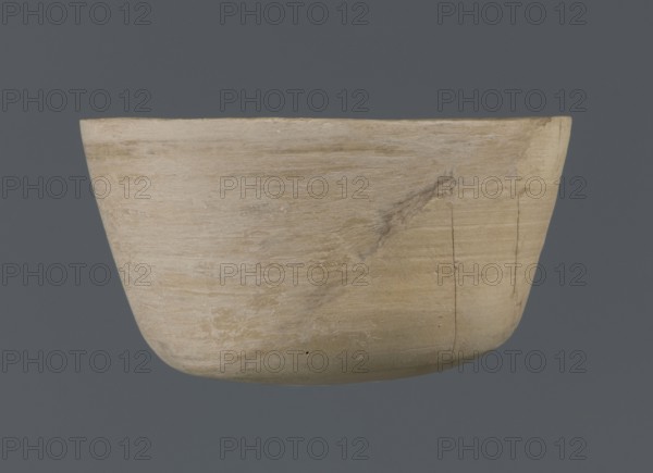 Bowl, unknown northern Mesopotamian workshop; Early Jazira III period ca. 2600–ca. 2350 BC / Early Dynastic III period ca. 2600–ca. 2350 BC; clay table vessel bowl with a flat oval bottom from which straight, slightly tilted walls rise in a short arc, terminating in a straight, not distinct rim; provenance: Al-Jazira Northern Mesopotamia, Khabur basin, Tell Jassa el Gharbi north-eastern Syria; vessel; height 7 cm, width 12.2 cm., A shallow rounded bowl with a smooth, worn surface shown against a solid dark gray background, the bowl displaying a range of beige and light tan tones with subtle streaks and patches of pale cream and faint brown, a few visible surface cracks and a darker smudge on one side, and the bowl's curved rim and gently rounded base clearly visible.