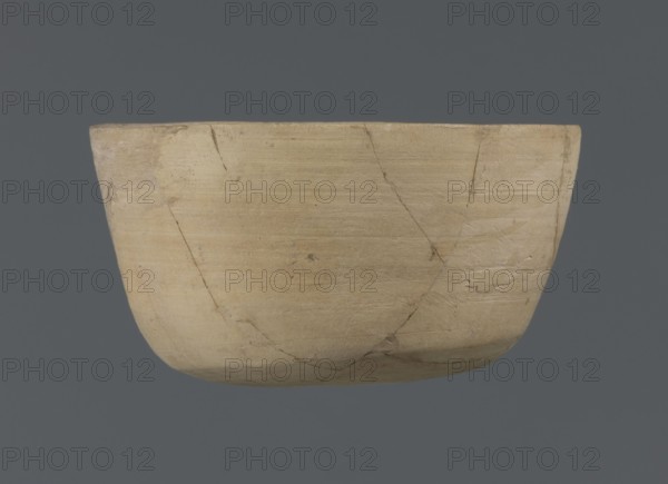 Bowl, unknown northern Mesopotamian workshop, Early Jazira III period ca. 2600–ca. 2350 BC, clay; table vessel. Bowl with a flat oval bottom from which straight, slightly tilted walls rise in a short arc, terminating in a straight, not distinct rim. Ca. 2600–ca. 2350 BC. From Tell Jassa el Gharbi North-Eastern Syria, Northern Mesopotamia Khabur basin. Vessel; height 7 cm, width 12.2 cm., A shallow rounded bowl shown from the side against a solid background, with a smooth, matte surface in shades of beige and tan and subtle darker hairline cracks and repair lines visible across its body, slight variations in tone with hints of pale cream and light brown, and a uniform gray background.