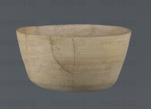 Bowl, unknown northern Mesopotamian workshop, Early Jazira III period ca. 2600–ca. 2350 BC. Clay table vessel — bowl with a flat oval bottom from which straight, slightly tilted walls rise in a short arc, terminating in a straight, not distinct rim. From Tell Jassa el Gharbi North-Eastern Syria, Northern Mesopotamia Khabur basin. Vessel; height 7 cm, width 12.2 cm. Materials and typology: clay, Early Dynastic III period; vice-less vessel; Al-Jazira region., A shallow, rounded bowl with a matte, slightly rough surface displayed against a uniform dark gray background, the bowl showing a light beige to pale tan color with subtle variations and a few darker beige streaks and smudges; the rim is thin and uneven, and the exterior surface bears visible horizontal tool marks and faint cracks where sections meet, while the interior appears a similar pale beige with slight discoloration and texture differences.