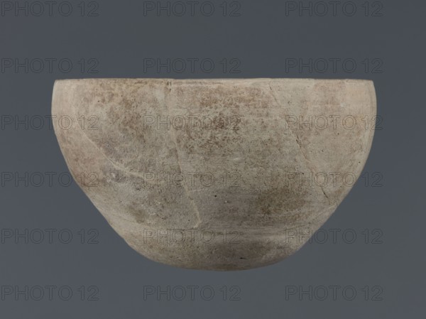 Bowl, unknown Northern Mesopotamian workshop, Early Jazira III period ca. 2600–ca. 2350 BC; Early Dynastic III period. Deep clay bowl with a rounded, slightly flattened, carelessly thickened bottom from which the walls emerge in an arched line. In the upper part the walls are slightly inclined toward the center. Straight, low rim distinguished by a carelessly engraved line on the outside. Vessel; height 8.7 cm, width 14.4 cm. Found at Tell Jassa el Gharbi, Northeastern Syria Al-Jazira, Khabur Basin, Northern Mesopotamia., A roughly hemispherical bowl with a rounded base and an open top, showing an uneven, textured surface with visible patches and subtle irregularities; the bowl displays a mix of beige, light brown, gray, and muted pink tones with some darker brown speckling and faint streaks, set against a solid dark gray background.