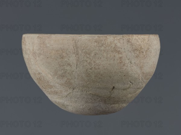 Bowl, unknown Northern Mesopotamian workshop, Early Jazira III period ca. 2600–ca. 2350 BC Early Dynastic III period. Deep clay bowl with a rounded, slightly flattened, carelessly thickened bottom from which the bowl walls emerge in an arched line. In the upper part the walls are slightly inclined toward the center. The straight, low rim is distinguished by a carelessly engraved line on the outside. Origin: Tell Jassa el Gharbi Northeastern Syria, Northern Mesopotamia Khabur Basin. Function: tableware vessel. Dimensions: height 8.7 cm, width 14.4 cm., A single rounded bowl-like object fills most of the frame, showing a rough, weathered surface with visible cracks and pitted texture; the object displays a palette of beige, light tan, pale gray, and hints of muted brown, with subtle variations and mottling across its curved exterior, set against a uniform dark gray background.