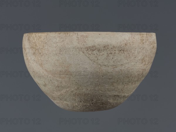 Bowl, unknown Northern Mesopotamian workshop, Early Jazira III period ca. 2600–ca. 2350 BC Early Dynastic III period, deep bowl with a rounded, slightly flattened, carelessly thickened bottom from which the bowl walls emerge in an arched line; in the upper part the walls are slightly inclined towards the center; straight low rim distinguished by a carelessly engraved line on the outside; clay vessel, tableware, vice-free vessel; Syria Tell Jassa el Gharbi, Northeastern Syria, Northern Mesopotamia Khabur Basin; height 8.7 cm, width 14.4 cm., A rounded bowl with a matte, textured surface in shades of beige, off-white, light tan, and muted gray, showing a few subtle darker brown and reddish-brown patches and speckles; the rim is slightly uneven and the body displays faint circular marks and hairline cracks on its coarse, weathered exterior against a solid dark gray background.