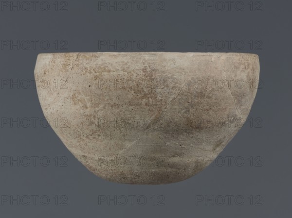 Bowl, unknown Northern Mesopotamian workshop, Early Jazira III period ca. 2600–ca. 2350 BC Early Dynastic III period. Deep bowl with a rounded, slightly flattened, carelessly thickened bottom, from which the bowl walls emerge in an arched line. In the upper part the walls are slightly inclined towards the center. The straight, low rim is distinguished by a carelessly engraved line on the outside. Clay vessel; tableware; vice-free vessel. From Tell Jassa el Gharbi, Northeastern Syria Khabur Basin, Northern Mesopotamia. Height 8.7 cm; width 14.4 cm., A rounded, bowl-shaped object with a rough, weathered surface showing a mix of beige, tan, light brown, and gray tones suspended against a solid dark gray background, with visible surface texture including areas of discoloration, fine cracks, and irregular patches suggesting uneven wear.