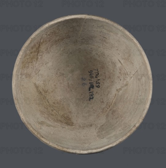 Bowl, unknown Northern Mesopotamian workshop, Early Jazira III period ca. 2600–ca. 2350 BC Early Dynastic III period. Deep bowl with a rounded, slightly flattened, carelessly thickened bottom from which the bowl walls emerge in an arched line. In the upper part the walls are slightly inclined towards the center. The straight, low rim is distinguished by a carelessly engraved line on the outside. Clay vessel; tableware. Origin: Northern Mesopotamia Khabur Basin, Al-Jazira; discovered at Tell Jassa el Gharbi Northeastern Syria. Height 8.7 cm, width 14.4 cm., A round shallow bowl viewed from above with a smooth inner surface showing a weathered, uneven texture; the bowl has a muted palette of beige, light tan, and pale brown with a few darker brown patches and small areas of grayish discoloration, and the rim appears slightly lighter than the interior.