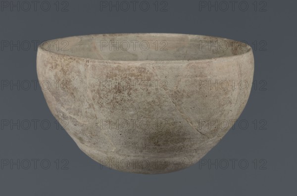 Bowl, unknown Northern Mesopotamian workshop, Early Jazira III period ca. 2600–ca. 2350 BC / Early Dynastic III period, deep clay bowl with a rounded, slightly flattened, carelessly thickened bottom from which the walls emerge in an arched line. Upper walls slightly inclined toward the center; straight low rim distinguished by a carelessly engraved line on the outside. Material: clay; function: vessel/tableware; type: bowl. Origin: Northern Mesopotamia Al-Jazira, Khabur Basin, Tell Jassa el Gharbi Northeastern Syria. Dimensions: height 8.7 cm, width 14.4 cm., A simple rounded bowl with a matte, textured surface showing a mix of gray, beige, tan, and muted brown tones, with subtle variations and mottled patches across its exterior and interior; the rim appears slightly irregular and worn, and the surface shows faint striations, small pitting, and a few hairline cracks, all set against a solid dark gray background.