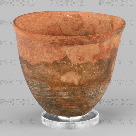 Mug, unknown Northern Mesopotamian workshop. Mug with slightly S-shaped walls and a small flat bottom with a convexity in the center. The rim is thickened in the form of a low, outwardly curled strip. Diagonal scratches are visible on the walls, leaving a technological trace of the vessel being polished. 1st half of the 3rd millennium BC; Early Jazira III period ca. 2600–ca. 2350 BC. Tell Jassa el Gharbi North-Eastern Syria, Northern Mesopotamia Khabur basin. Clay vessel, tableware; height 8.2 cm, width 9.5 cm, weight 156 g., A rounded ceramic vessel resting on a clear circular stand, showing a textured surface with areas of reddish-brown, terracotta, tan, olive green, and darker brown; the upper rim is slightly flared and the interior shares the reddish-brown and tan tones; the exterior displays a mottled pattern with irregular patches and streaks, and the lower portion has horizontal striations and darker brown banding; the clear stand is glossy and colorless, and the background is a...