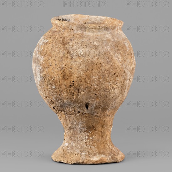 Beaker, unknown Northern Mesopotamian workshop; hand-made, asymmetrical miniature cup on a stem terminating in a flat, round foot. The cup's bulbous bowl is inclined inwards and terminates with a diagonally protruding, low rim. Clay vessel; miniature cup on stem. 1st half of the 3rd millennium BC; Early Jazira III period Early Dynastic III; ca. 2600–ca. 2350 BC. Origin: Northern Mesopotamia Khabur basin; discovered at Tell Jassa el Gharbi north-eastern Syria. Weight: 256 g., A weathered pottery vessel with a rounded body narrowing into a flared foot, shown against a plain gray background; the vessel surface appears rough and pitted with an uneven texture and small holes, exhibiting shades of tan, brown, beige, and hints of off-white residues; the rim is slightly irregular and the overall shape has an organic, aged appearance.