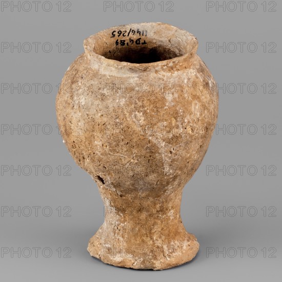 Beaker, unknown Northern Mesopotamian workshop; hand-made, asymmetrical miniature cup on a stem terminating in a flat, round foot. The cup's bulbous bowl is inclined inwards and terminates with a diagonally protruding, low rim. Clay vessel; miniature cup on stem. 1st half of the 3rd millennium BC — Early Jazira III period Early Dynastic III; ca. 2600–ca. 2350 BC. Tell Jassa el Gharbi North-Eastern Syria; Northern Mesopotamia Khabur basin. Weight: 256 g., A roughly rounded ceramic vessel with a narrow flared neck and a pedestal base fills the image, showing a weathered, textured surface in shades of tan, beige, light brown, and off-white with patches of darker brown and subtle grayish tones; the vessel has irregularities and small pits in the clay, a slightly uneven rim, a few small chips and rough edges, and a visible hollow interior that appears darker than the exterior; the background is a uniform medium gray.