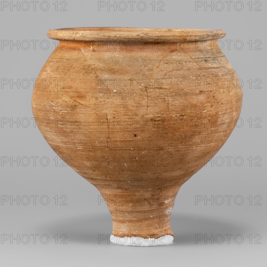 Beaker, unknown Northern Mesopotamian workshop; stemmed beaker, clay; Beaker with a bulging bowl, with an upturned flat and narrow rim, set on a low cylindrical stem terminating in a small foot damaged. Horizontal traces of polishing on the surface. 1st half of the 3rd millennium BC; Early Jazira III period ca. 2600–ca. 2350 BC. Tell Jassa el Gharbi north-eastern Syria, Northern Mesopotamia Khabur Basin. Vessel; height 11.5 cm, width 11.6 cm, weight 264 g., A rounded pottery vessel with a narrow base and flaring rim sits against a plain background, its surface textured with visible horizontal striations and small rough patches; the vessel is primarily warm brown and tan with subtle variations in tone and a few lighter flecks, and there is a small area of white material at the base.